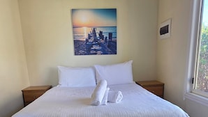 A neatly made bed with white linens and towels, a cityscape painting on the wall, and a window with a view of greenery. - BIG4 Kingston Foreshore Holiday Park (Kingston SE)