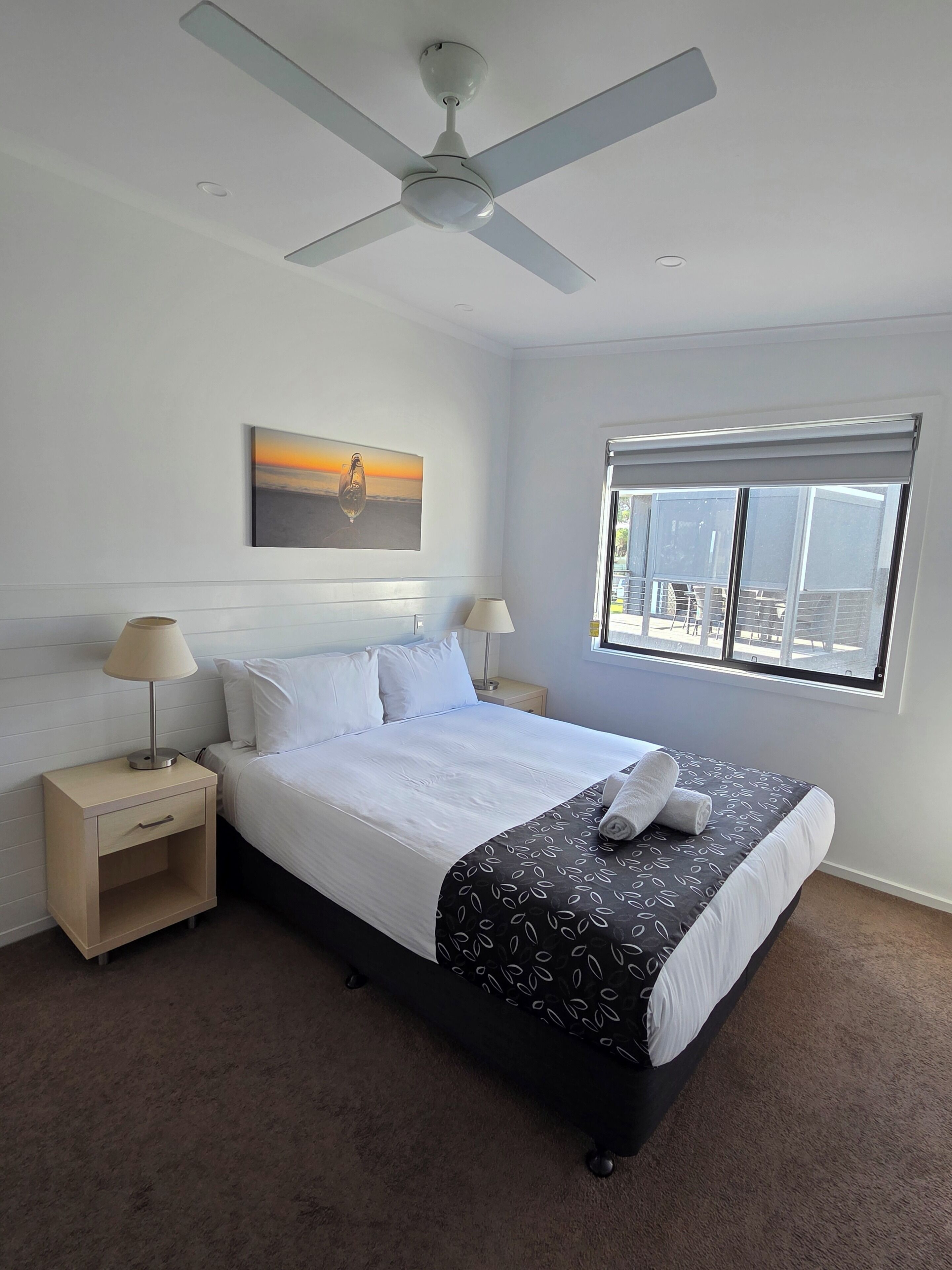 A hotel room with a bed, a ceiling fan, a window with a view, and a bedside table with a lamp.