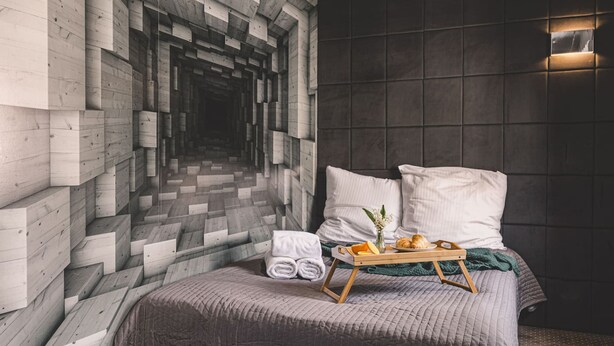 A bedroom with a unique wall design, a bed with a tray of breakfast items, and a neatly made bed with a rolled towel.