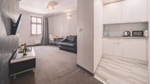 A modern living room with a sofa, a small table, and a TV. There is a kitchen area with cabinets and a microwave.