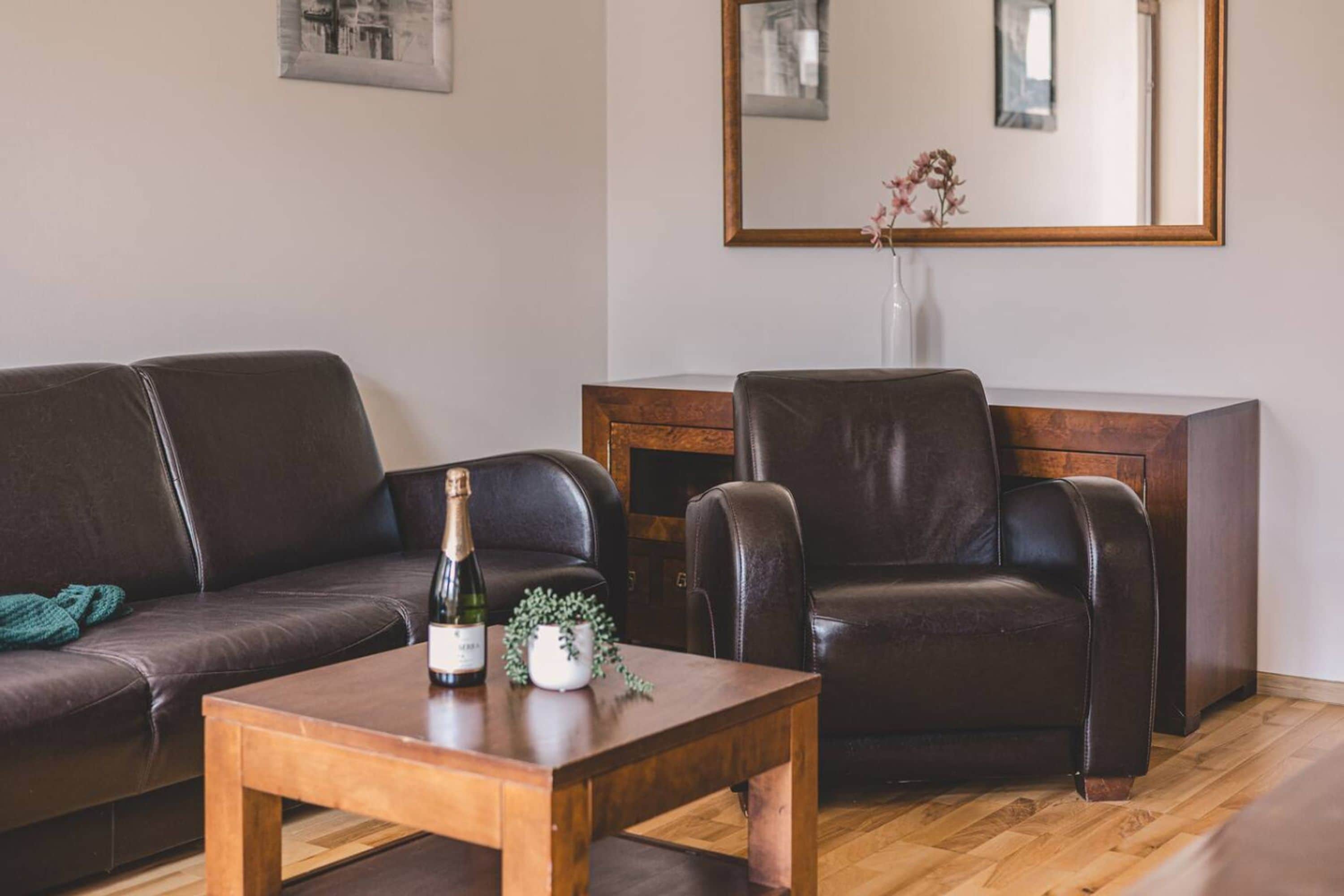 A living room with a brown leather sofa, a wooden coffee table, and a leather armchair. A bottle and a small plant are on the table.