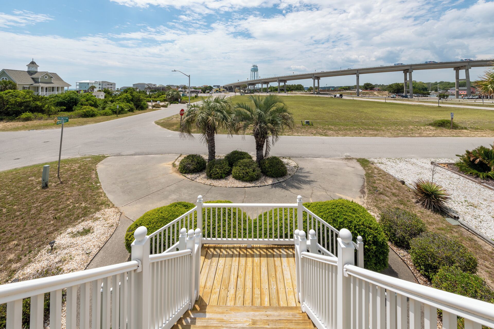 A wooden deck with white railings leading to a palm tree surrounded by bushes.