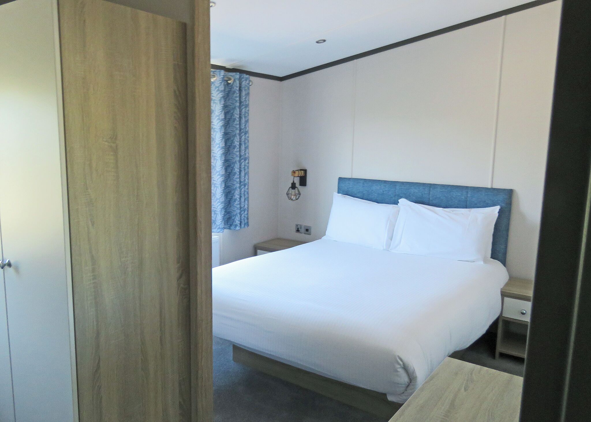 A hotel room with a bed, a nightstand, a wall-mounted lamp, and a window with curtains.