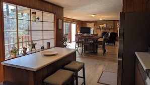Une cuisine avec des lambris en bois, une grande fenêtre avec des stores, un coin repas avec une table et des chaises, et un réfrigérateur.