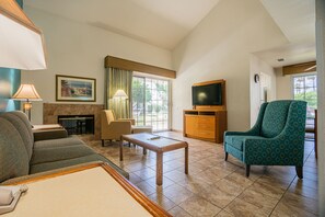 Salón de hotel con sofá, sillón, mesa de centro, chimenea, televisor y un amplio ventanal con vistas al exterior.