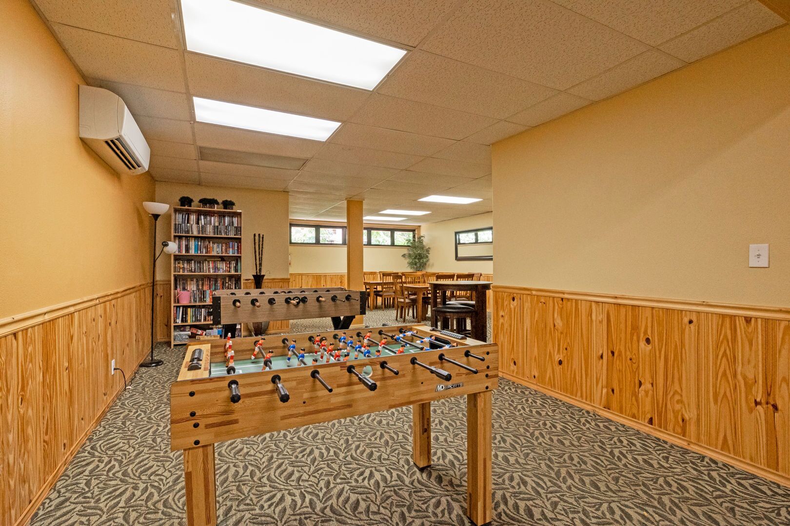 A room with a foosball table, a bookshelf, and a dining area.