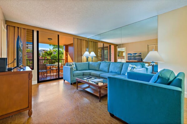 A modern hotel room with a large sofa, a coffee table, and a TV on a wooden cabinet. There is a balcony with a view of palm trees and a building.