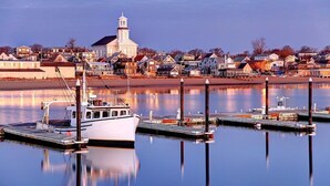 A marina with a white boat docked, a church with a steeple, and a row of houses along the water. - 1BR Condo w/ Kitchenette & Balcony | Pools Access (South Yarmouth)