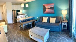 A modern living room with a sofa, coffee table, and a dining area with a bench. - 1BR Condo w/ Kitchenette & Balcony | Pools Access (South Yarmouth)