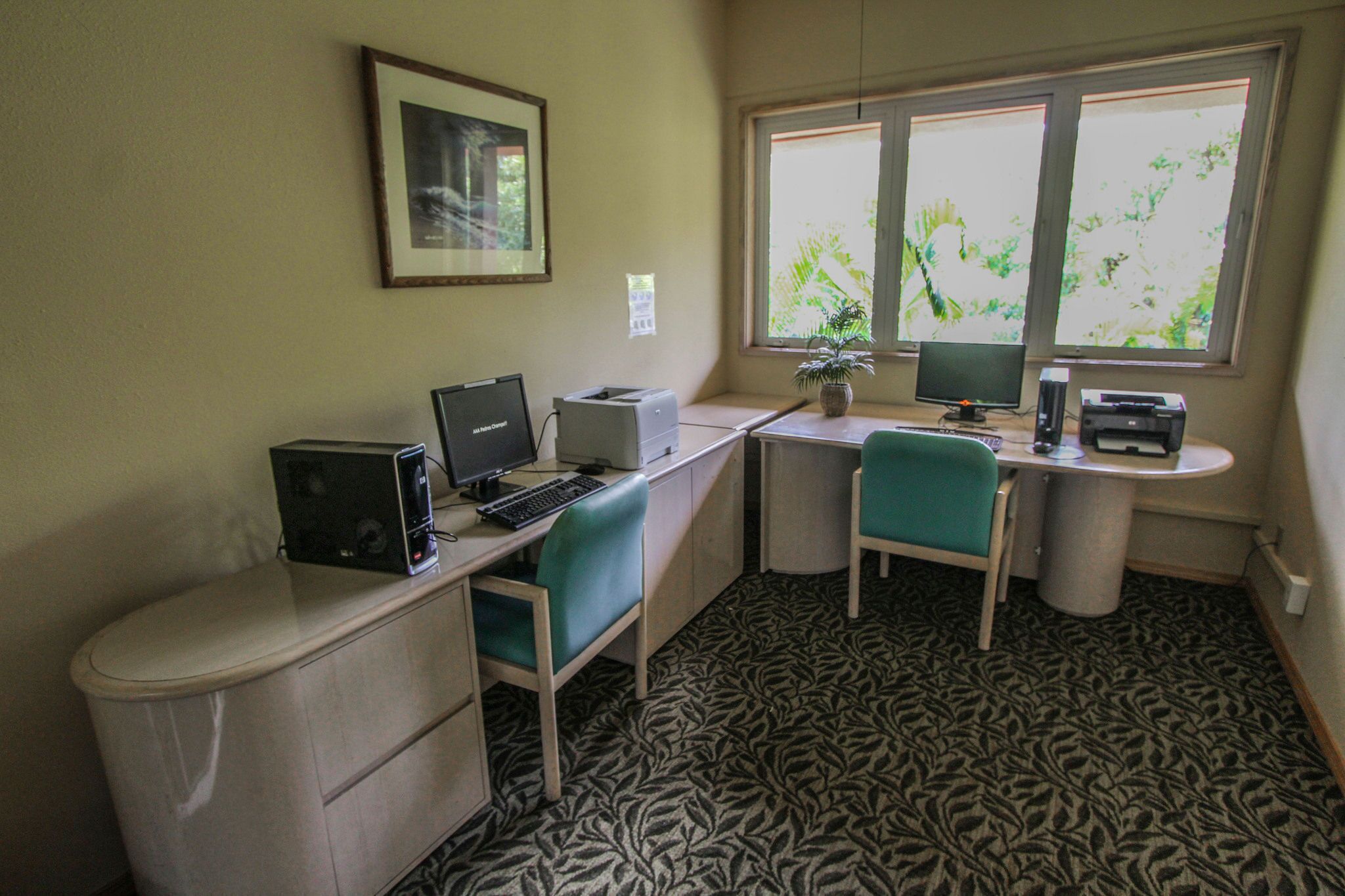 Un petit bureau avec un bureau, deux chaises, une imprimante, un ordinateur et un tableau encadré accroché au mur.