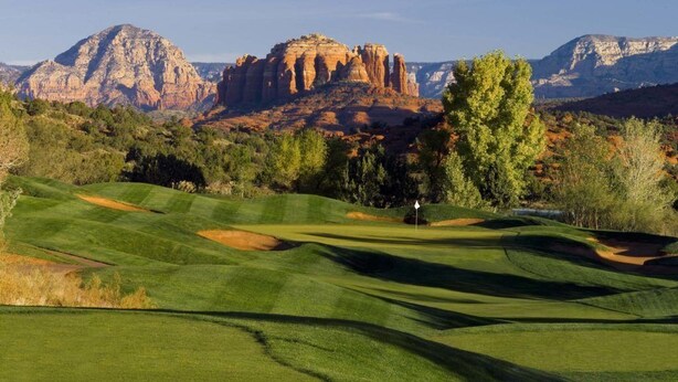 A golf course with a flagstick, surrounded by green grass, trees, and red rock formations in the background.