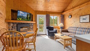 L’intérieur d’un chalet accueillant, avec des murs en bois, une cheminée en pierre, un téléviseur, une table à manger avec des chaises, un canapé et une fenêtre avec des rideaux.