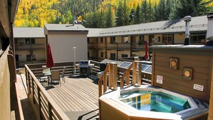 Una terraza de madera con un jacuzzi, mobiliario exterior y vistas a árboles otoñales.