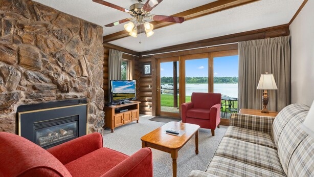 A cozy living room with a stone wall, a fireplace, a television, a red armchair, a plaid sofa, and a view of the outdoors through large windows. - Point Condos 2BR w/ Lake Views & Fireplace (Breezy Point)