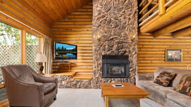 Interior de una cabaña de troncos con chimenea de piedra, un televisor y una zona de estar acogedora con sofá y sillón.