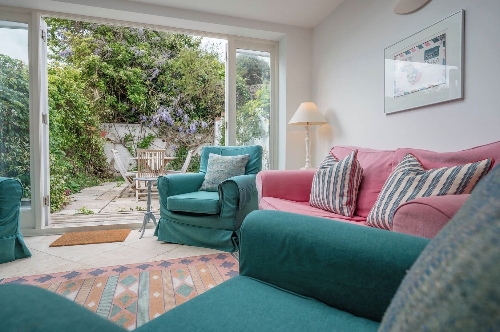 Un salon chaleureux avec des canapés turquoise et roses, un tapis à motifs et une vue sur un jardin luxuriant, visible à travers une porte vitrée.