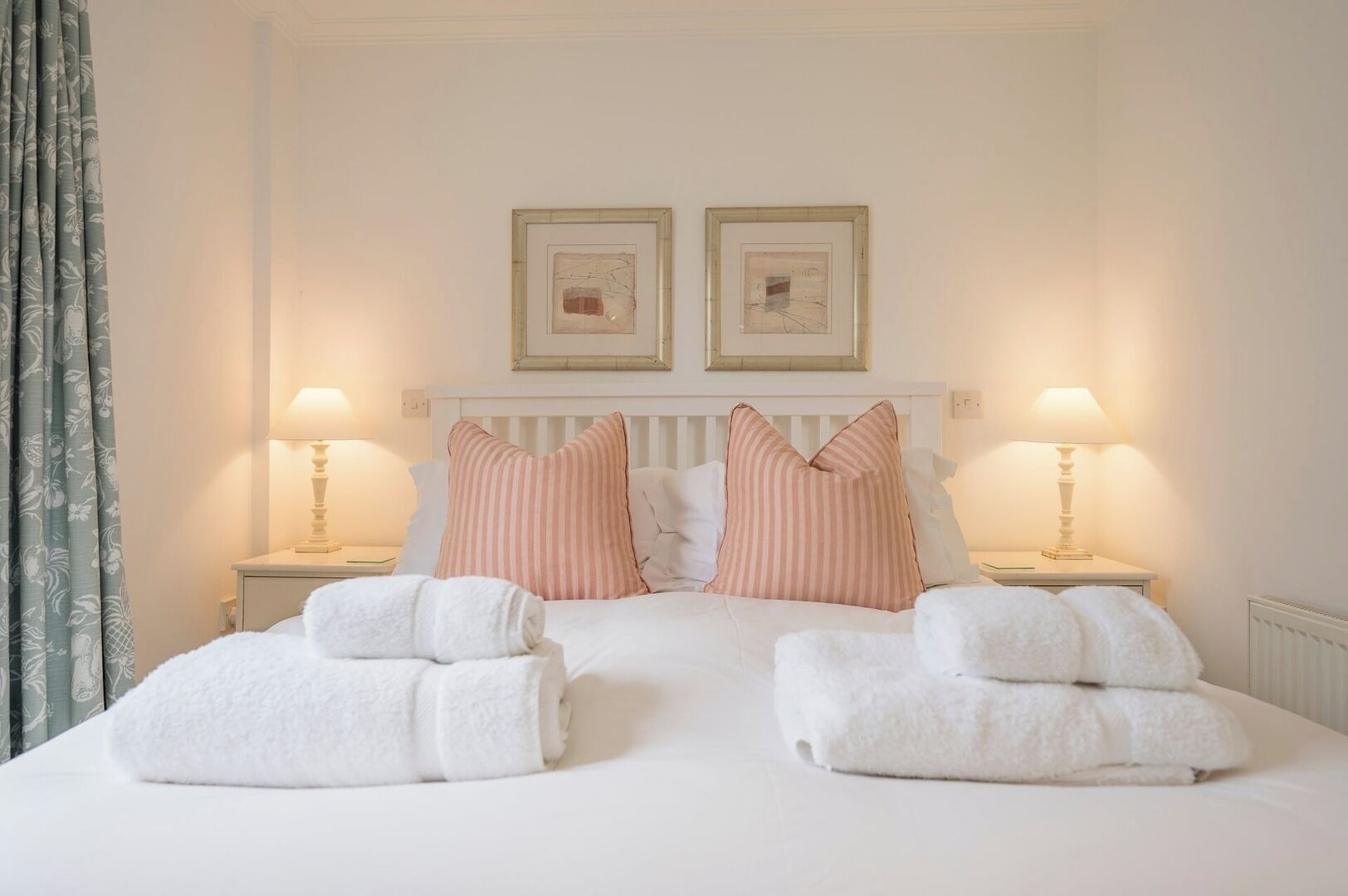 A double bed with white linens, two striped pillows, and two white towels. There are two bedside tables with lamps and framed pictures above the bed.