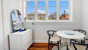 A room with a white dresser, a round table with four chairs, a large window with a city view, and a mirror on the wall.