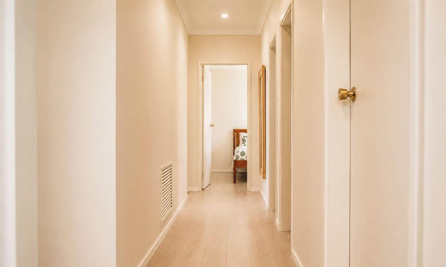 A hallway with a door at the end, a radiator on the wall, and a bed with a patterned bedspread in the background.