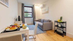 A modern living room with a dining area, a grey sofa, a black shelf with fruits, and a window with curtains. - Cozy & Bright Flat by Host Wise (Guimarães)