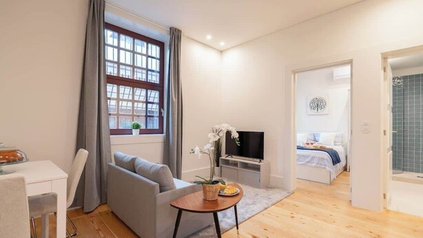 A modern living room with a grey sofa, a small wooden table with a plant, a TV, and a dining area with a white table and chairs. - Modern & Cozy Flat by HostWise (Guimarães)