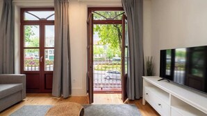 A modern living room with a sofa, a TV on a white cabinet, and a view of the street through a glass door.
