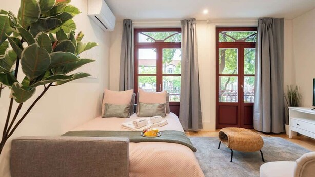 A bedroom with a bed, a bedside table, a chair, a plant, and a window with curtains.