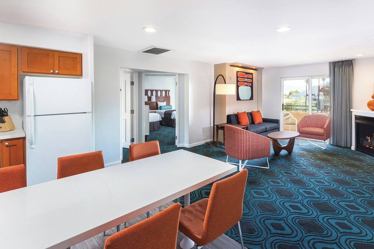 A modern hotel room with a dining area, a living room with a sofa and armchairs, a kitchen with wooden cabinets, and a fireplace.
