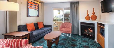 A hotel room with a sofa, armchair, fireplace, television, and a patterned carpet.