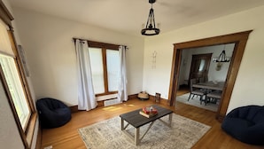A spacious living room with wooden flooring, a central coffee table, two bean bags, a rug, and a dining area visible through an open door. - Cozy Farmhouse Getaway 10 min from Lost Island and Casino (La Porte City)