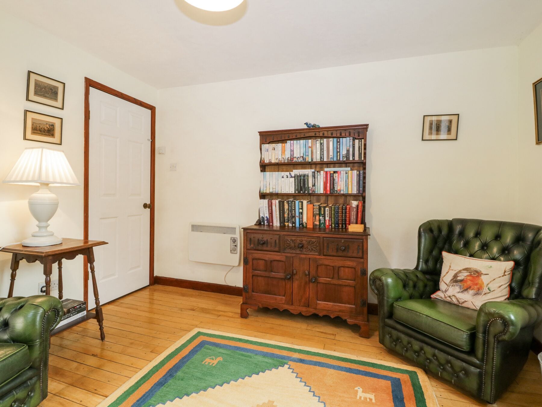 A room with a wooden bookshelf, a green leather armchair, a small table with a lamp, and a patterned rug.