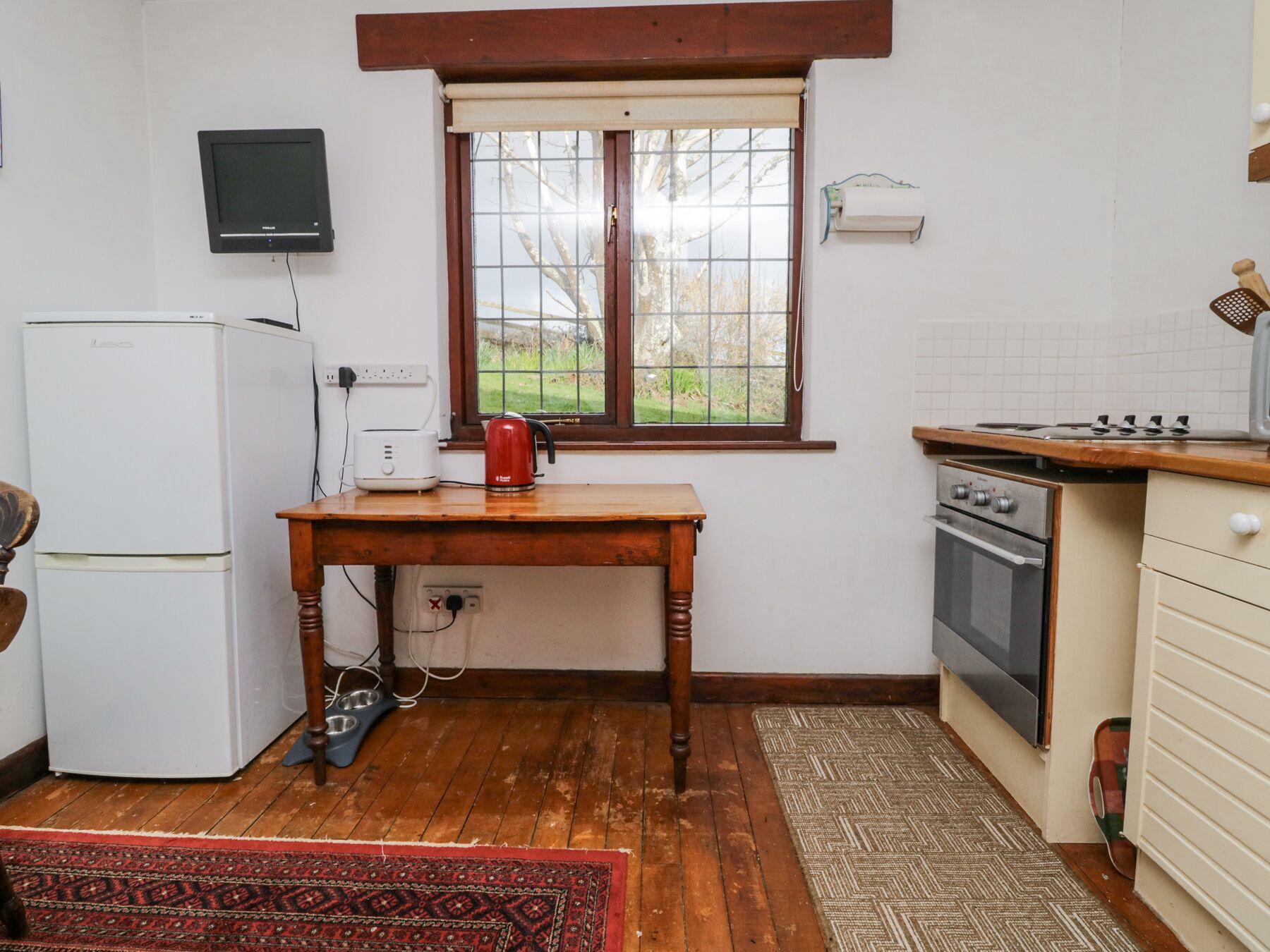Eine kleine Küche mit einem Kühlschrank, einem Toaster, einem Wasserkocher, einem Fenster mit Blick auf Bäume, einem Herd und einem Spülbecken.
