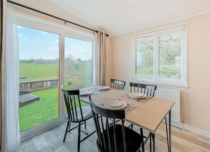 A dining area with a table set for four, large windows with curtains, and a view of a green landscape.