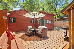 Una terraza de madera con una mesa y sillas, una sombrilla cubierta y un edificio con puerta y ventanas verdes.