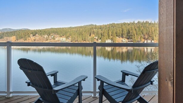 A wooden deck with two Adirondack chairs overlooking a calm lake, surrounded by a forested area. - The Boathouse Large Modern Renovated Waterfront Home with Decks and Docks (Coeur d'Alene)