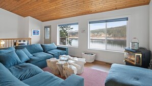 A cozy living room with a wooden ceiling, large windows with a lake view, a blue sofa, and a vintage stove. - The Boathouse Large Modern Renovated Waterfront Home with Decks and Docks (Coeur d'Alene)