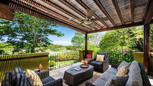 Un patio al aire libre con mobiliario de mimbre, un ventilador de techo y vistas a un jardín exuberante.