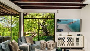 Una sala de estar moderna con un televisor grande, asientos cómodos y vistas a una playa tropical a través de amplios ventanales.