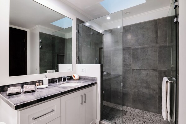 Un baño moderno con un espejo grande, encimera de mármol, ducha con mampara de vidrio y pared revestida de baldosas oscuras.