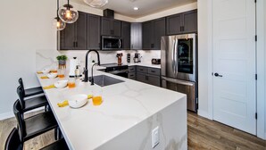 A modern kitchen with dark cabinets, a white marble island, stainless steel appliances, and black bar stools. - Villa Solara – Ultimate Backyard & Gameroom (Lake Havasu City)