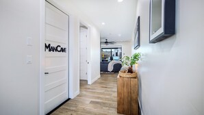 A hallway with a wooden floor, a white door labeled "MANCAVE," a wooden side table with a plant, and a ceiling fan. - Villa Solara – Ultimate Backyard & Gameroom (Lake Havasu City)