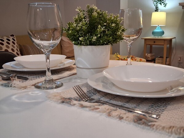 A dining table set with white plates, cutlery, and two wine glasses, accompanied by a potted plant.