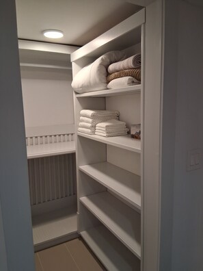 A walk-in closet with shelves for folded towels and blankets, a radiator below, and a light fixture on the ceiling.