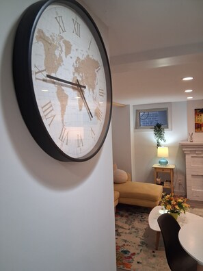A wall clock with a world map, a sofa, a small table with a lamp, and a window with a view of the outside.