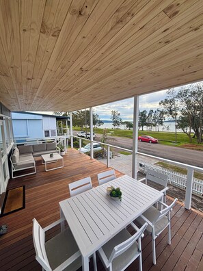 Ein überdachter Außenbereich mit Holzterrasse, weißen Möbeln und Blick auf eine Straße und ein Gewässer.