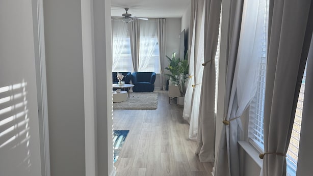 A hallway with a light-colored wooden floor, a tufted bench, and a view into a living room with a blue sofa and a ceiling fan. - Charming 3-bedroom townhome with loft, WiFi and AC in peaceful Lawrenceville (Lawrenceville)
