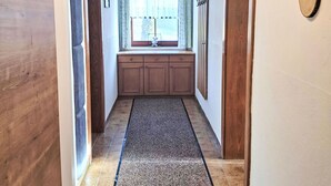 Ein schmales Entree mit Holzdielen, einem dunklen, gemusterten Teppich, einem Fenster mit durchsichtigen Vorhängen und einem Holzkorpus mit Waschbecken.