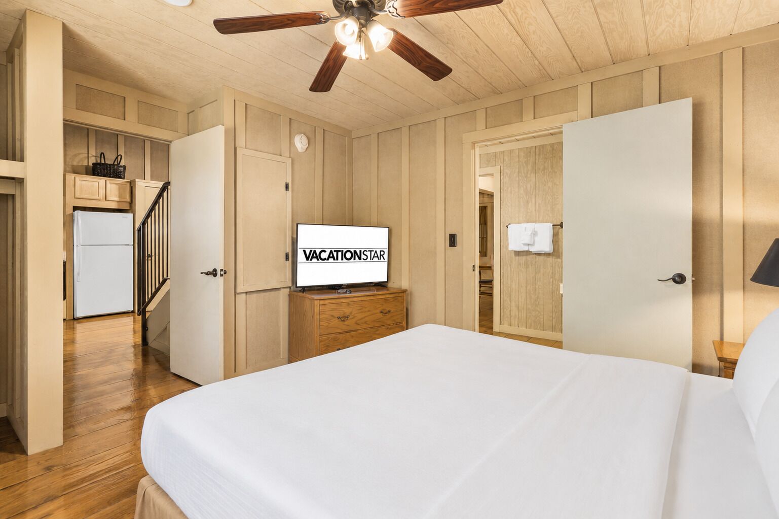 Habitación de hotel con cama, televisor mostrando 'VACATIONSTAR', ventilador de techo, paredes de madera y una cocina al fondo.