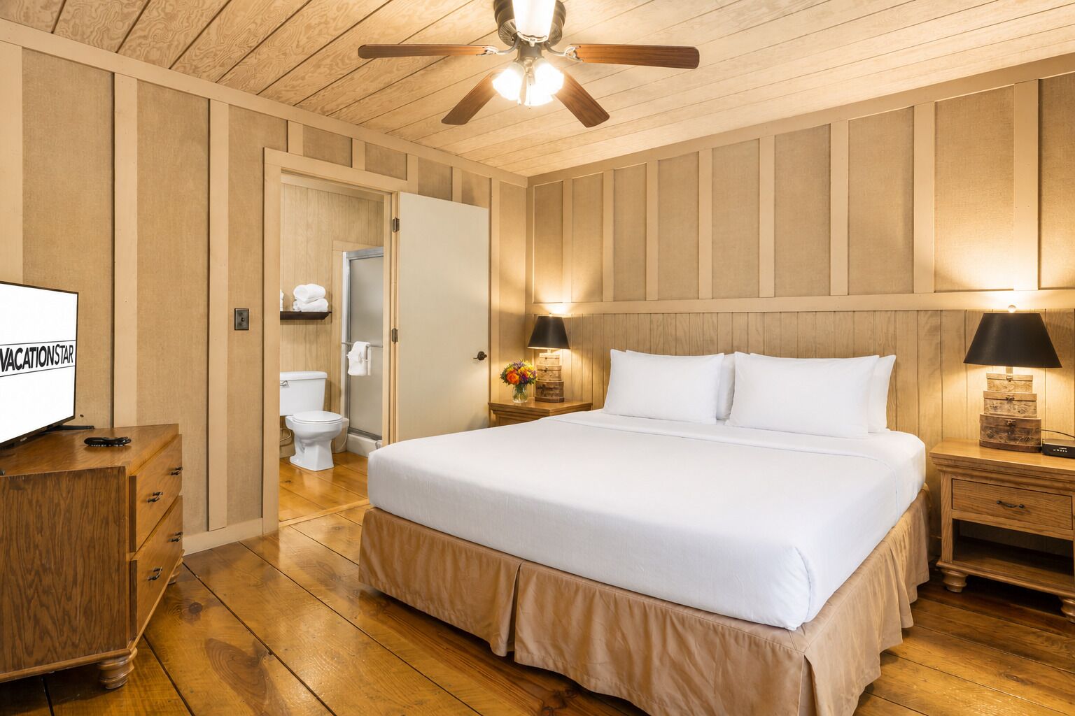 A bedroom with a bed, wooden walls, a ceiling fan, a TV, and a bathroom with a toilet and shower.