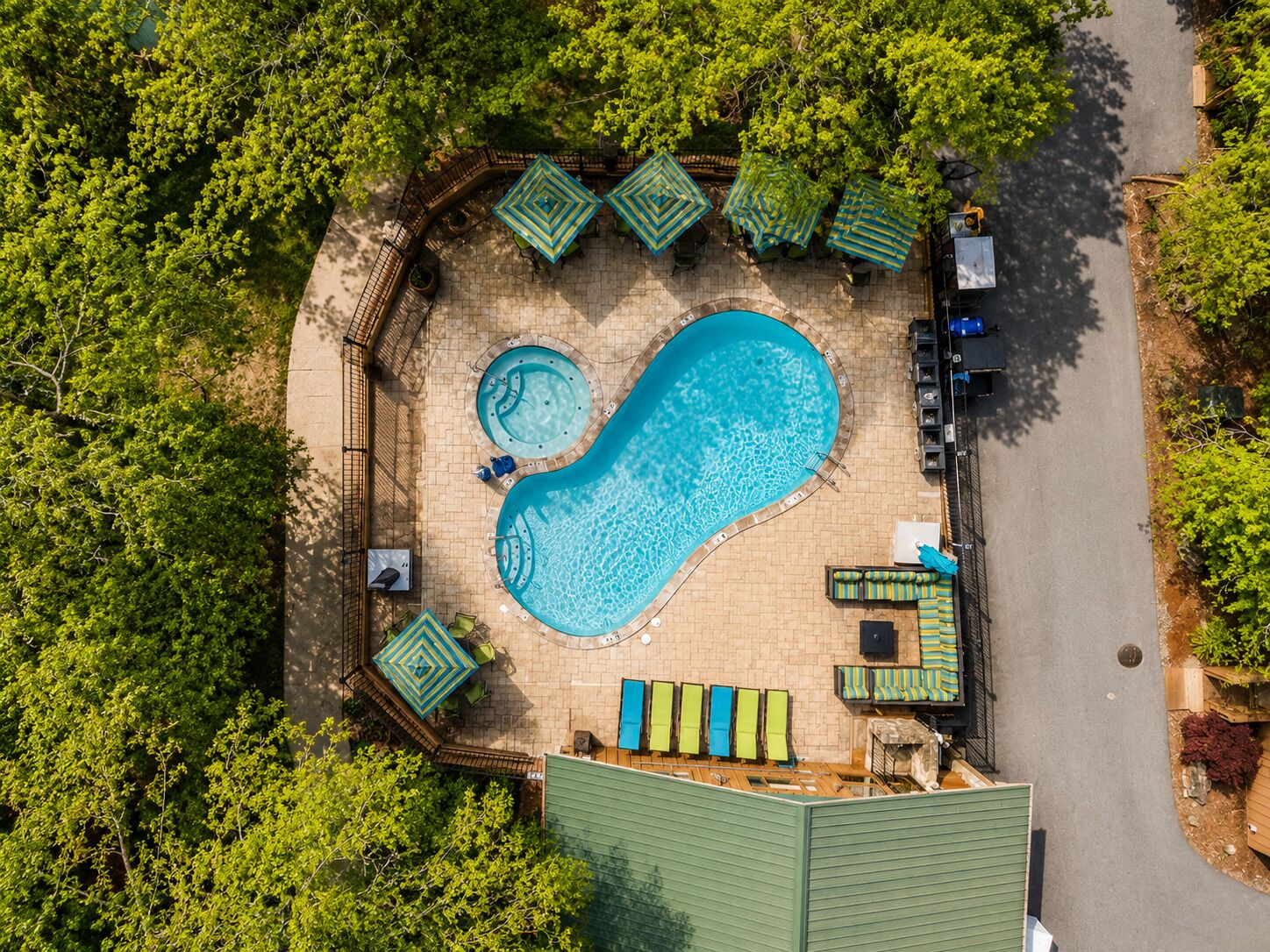 An aerial view of a swimming pool with two hot tubs, surrounded by lounge chairs and umbrellas, set in a landscaped area with trees.